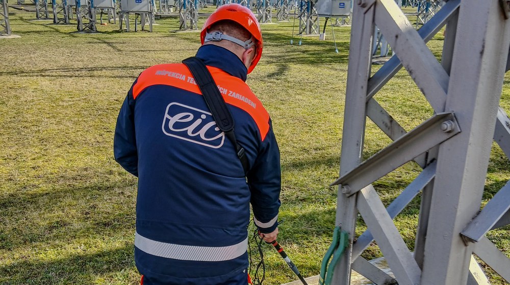 Inšpektor EIC v prilbe a pracovnom odeve s logom spoločnosti meria uzemnenie oceľovej konštrukcie priamo v elektrickej stanici.