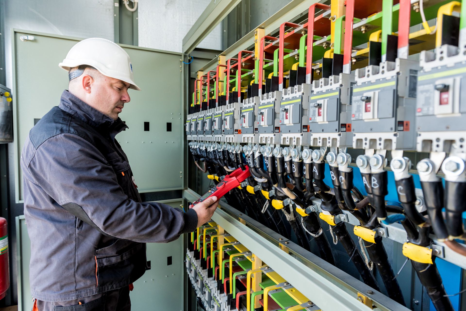 Revízny technik v bielej prilbe kontroluje kliešťovým meracím prístrojom istiace prvky vo veľkom priemyselnom rozvádzači.